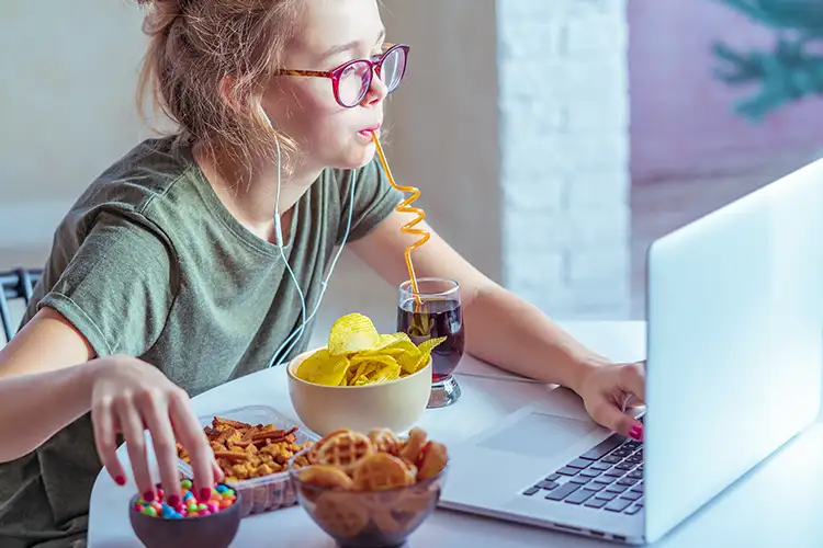 Junge Frau sitzt vor dem Computer und isst Fast Food – symbolisiert stressigen Studentenalltag, ungesunde Ernährung und digitalen Lebensstil