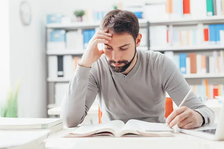 Junger Mann sitzt nachdenklich mit der Hand am Kopf zwischen Büchern und Notizen – sinnbildlich für Prüfungsstress