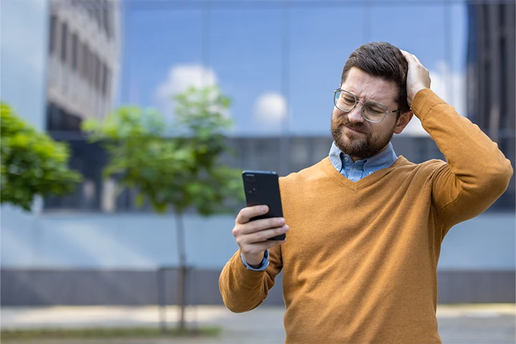 Ein Mann steht im Freien, blickt sorgenvoll auf sein Handy und hält eine Hand auf den Kopf.